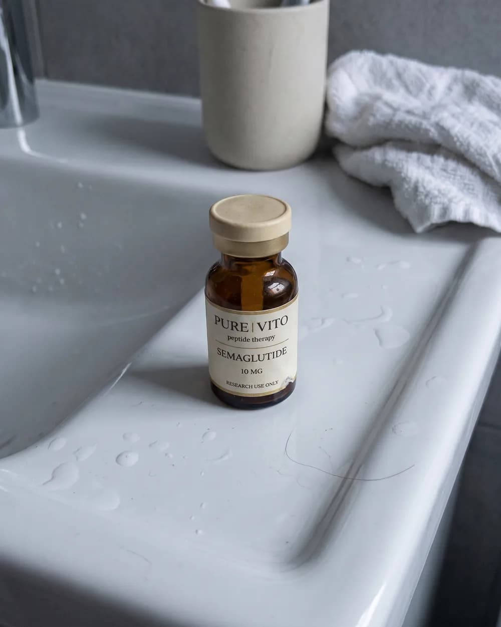 A Semaglutide vial standing on a wet bathroom basin ledge, beside a toothbrush cup and a crumpled towel.