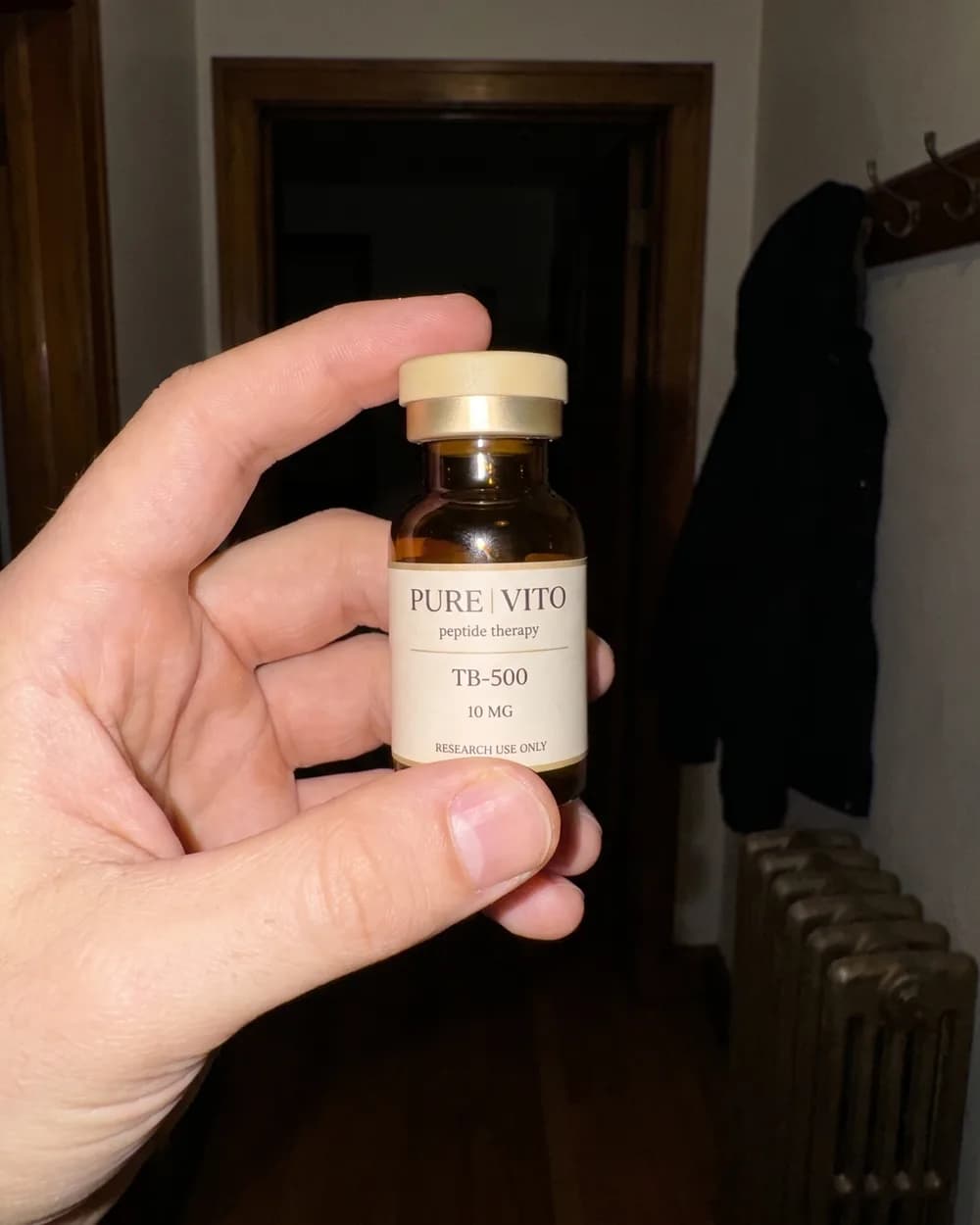 A hand holding up a TB-500 vial in a hallway, lit by the phone's flash.
