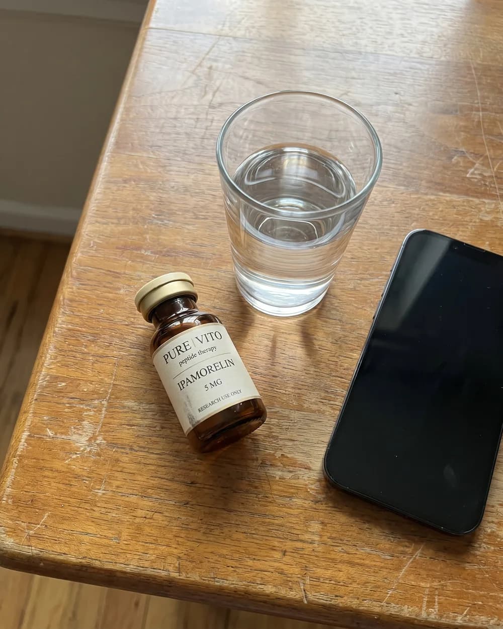 An Ipamorelin vial lying on its side on a scratched wooden table, beside a glass of water and a phone.