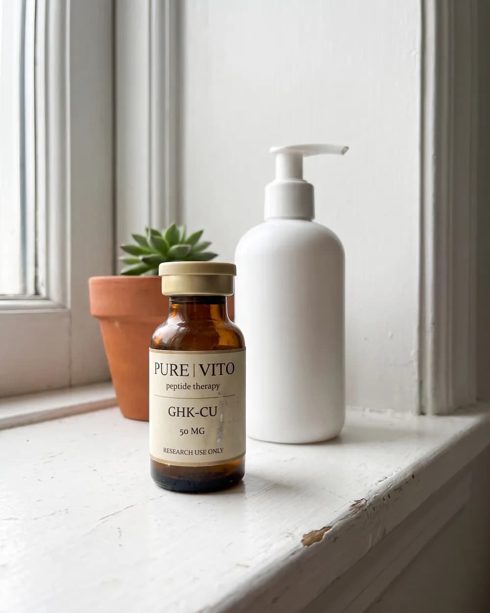 A GHK-Cu vial on a white windowsill beside a plain pump bottle and a small potted plant.