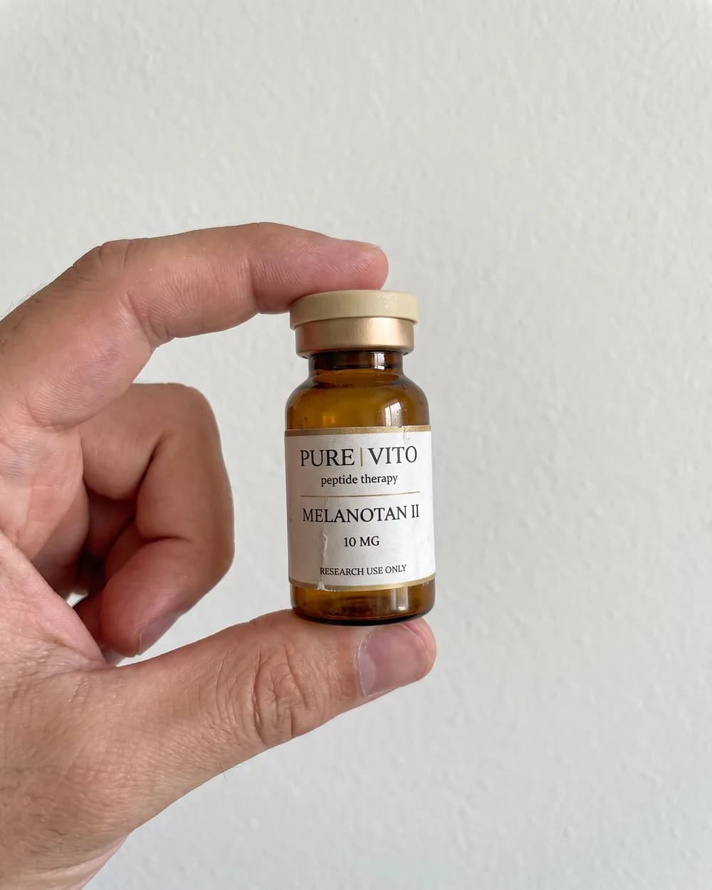 A Melanotan II vial held up between finger and thumb in front of a plain empty wall.