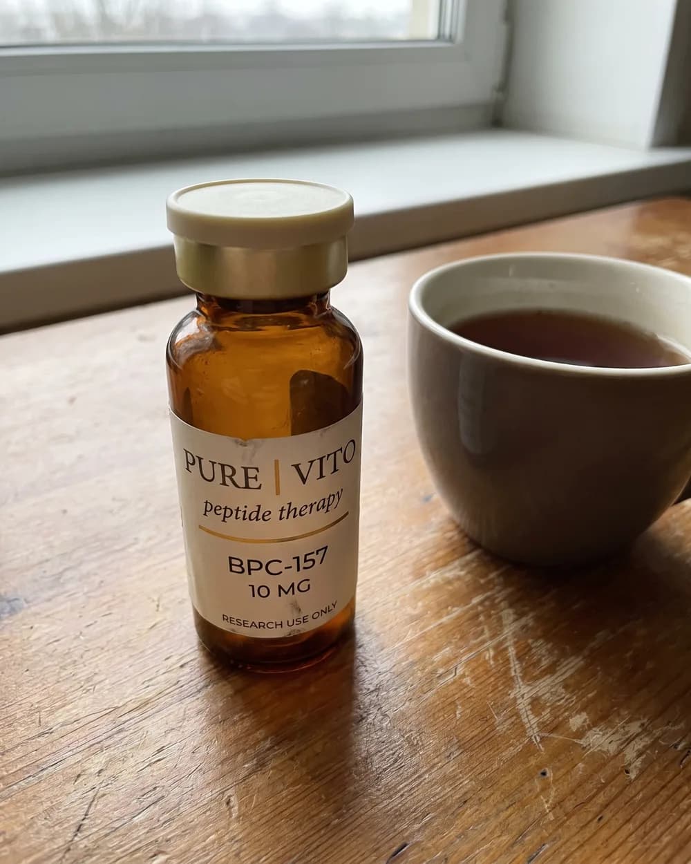 A BPC-157 vial standing alone on a wooden table by a window, next to a half-full mug of tea.