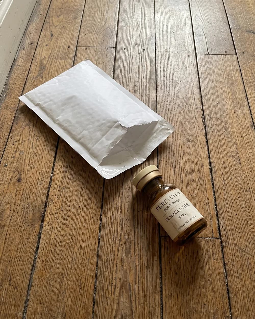 A blank padded mailer torn open on hallway floorboards, with a Semaglutide vial lying on its side beside it.